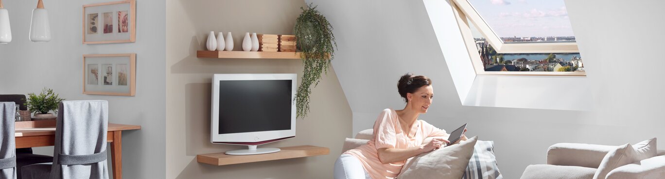 Ein Wohnzimmer mit einem Dachfenster. Eine Frau lächelt und sitzt auf einem Sofa unter dem Dachfenster.